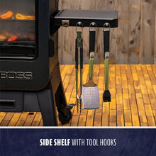 Black metal grill side shelf with three tool hooks holding tongs, a spatula, and a basting brush.