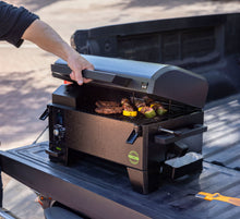 Compact black portable pellet grill with digital controls, open lid, grilling meat and veggies, set on a truck bed.