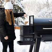 Black Sportsman pellet grill with woman adding pellets in snowy setting.