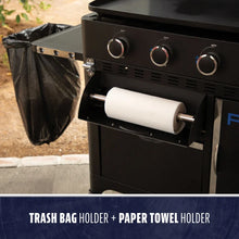 Black grill with attached trash bag holder and paper towel holder for easy outdoor cleanup and convenience.